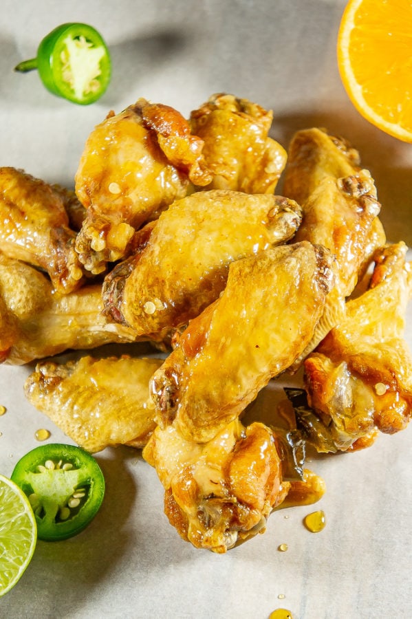 a pile of tequila lime wings on a piece of parchment paper with oranges, sliced jalapeno and lime next to it