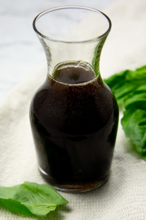 a small glass carafe of basil balsamic vinaigrette on a beige towel surrounded by fresh basil leaves
