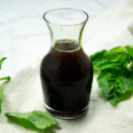 a small glass carafe of basil balsamic vinaigrette on a beige towel surrounded by fresh basil leaves