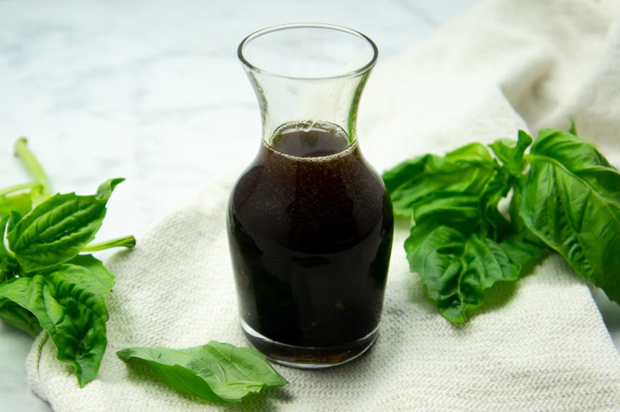 a small glass carafe of basil balsamic vinaigrette on a beige towel surrounded by fresh basil leaves