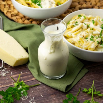 a glass carafe of white cheese sauce in front of a bowl of pasta