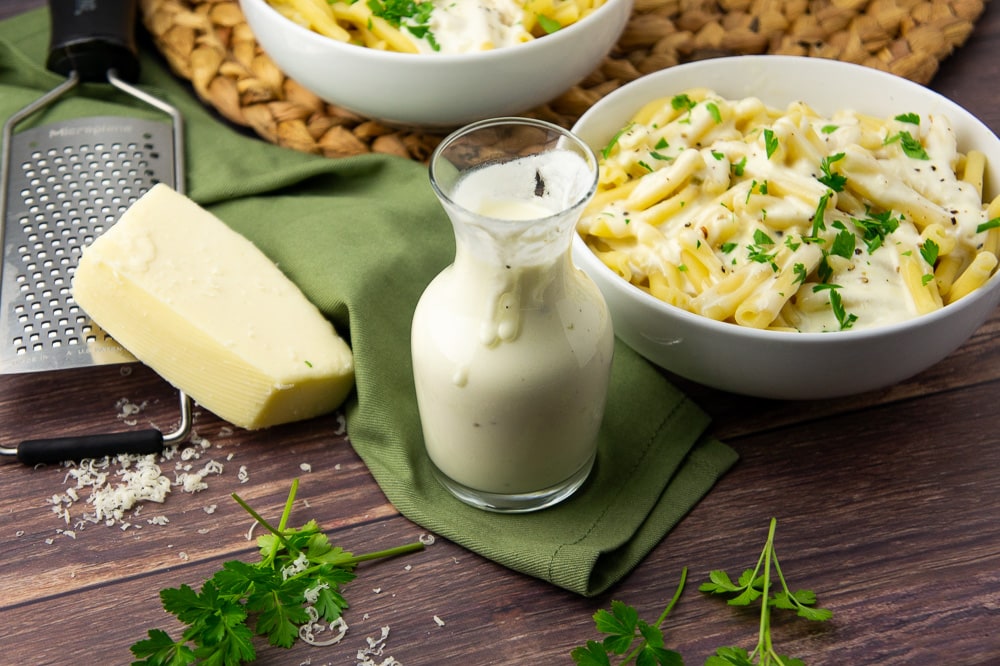 a glass carafe of white cheese sauce in front of a bowl of pasta