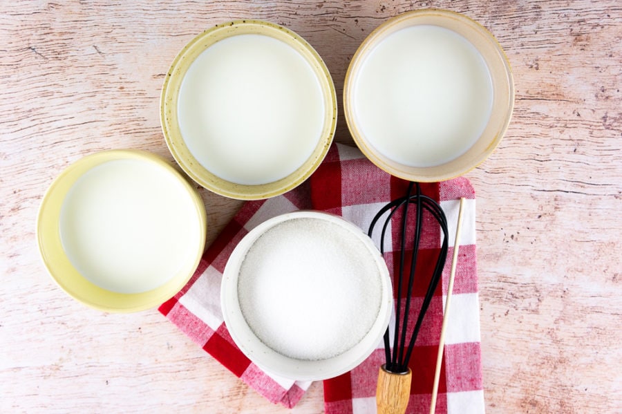 three bowls of milk, and 1 bowl of white sugar, a whisk and a wooden skewer on a red checked napkin