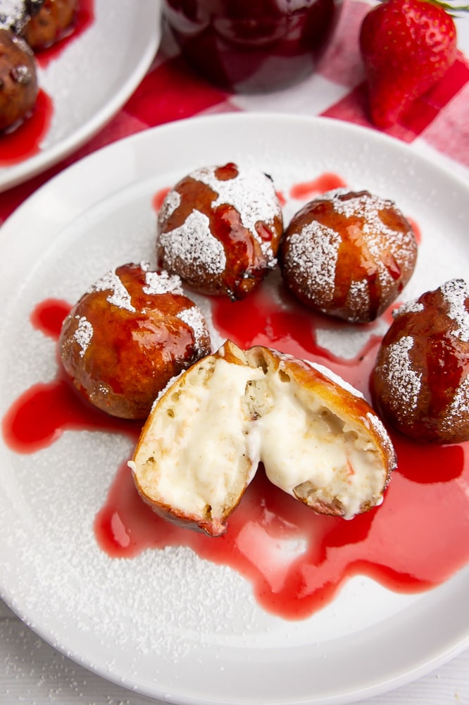 fried cheesecake bites with strawberry syrup on a white plate with one bite cut open and oozing creamy cheesecake