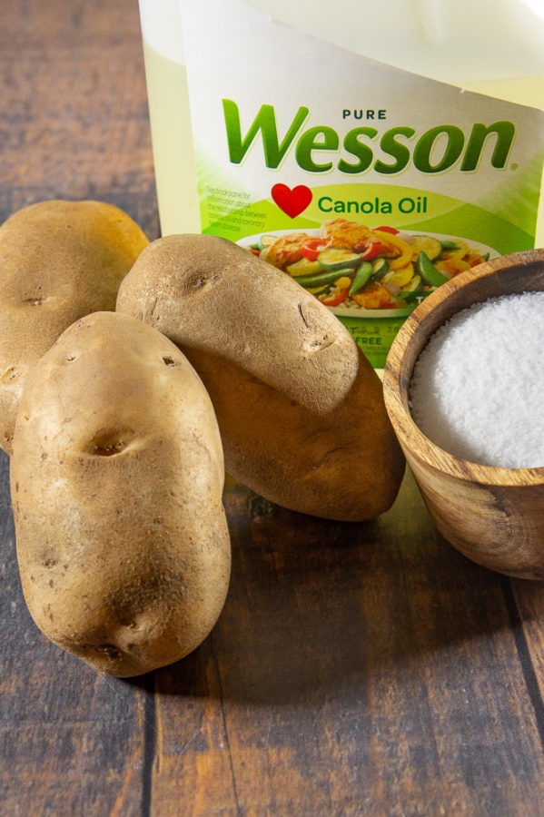 a pile of potatoes next to a bowl of salt and jug of canola oil, on a wooden table