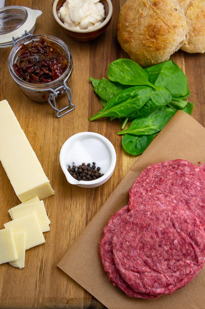 a wooden cutting board with four burger patties, white cheddar cheese, spinach, salt, pepper, and a jar of bacon jam