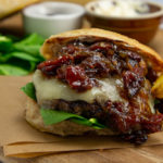 a wooden cutting board with a bacon onion jam burger, fries, a bowl of mayo and fresh spinach