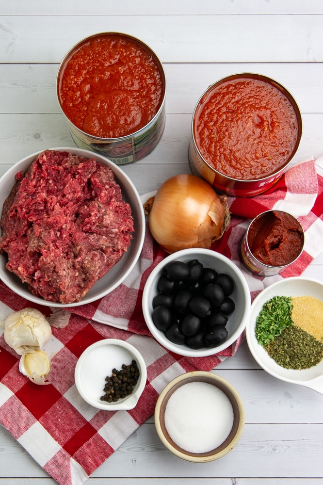 A bowl of ground beef, an onion, a bowl of olives, a bowl of dried herbs, a bowl of salt, and several cans of tomato sauce on a white wooden table