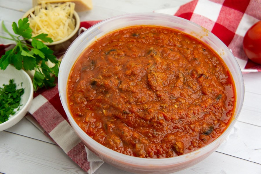 a large bowl of spaghetti sauce on a white wooden table with a red and white checked napkin beside parsley and Parmesan cheese. 