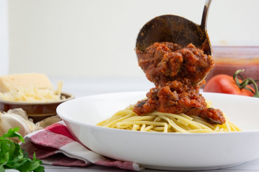a metal ladle pouring spaghetti sauce over a bowl of cooked spaghetti noodles. 