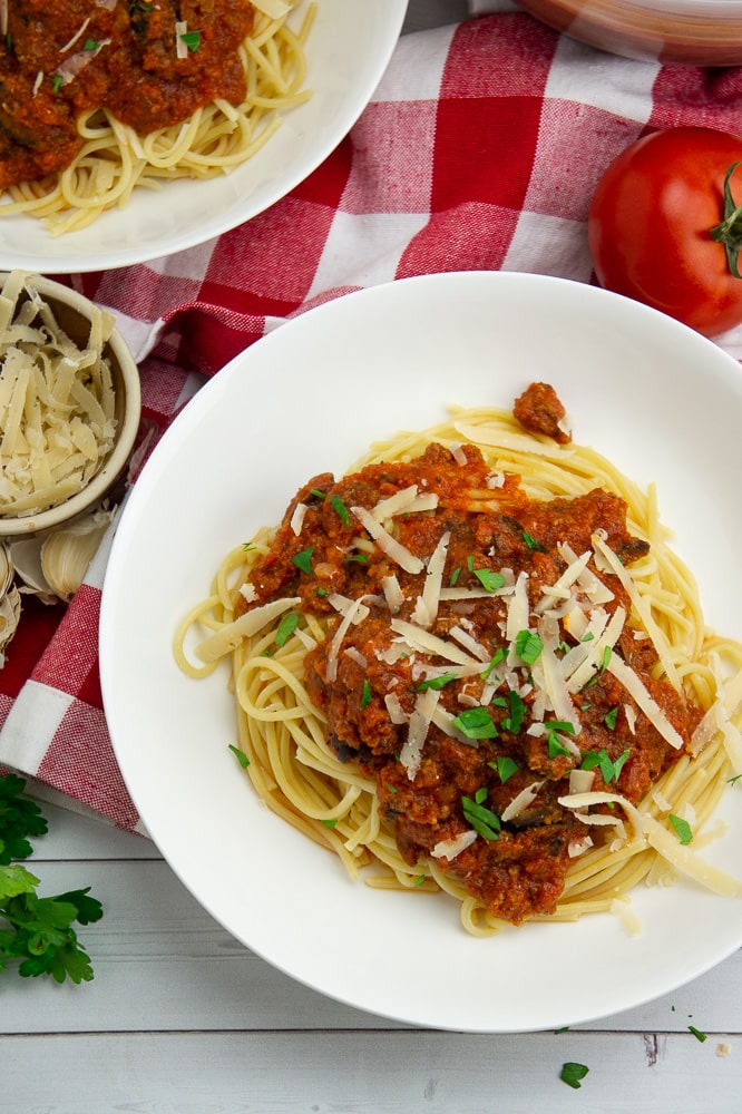 a white bowl with spaghetti noodles and meat sauce with fresh parmesan and parsley on top, beside a red and white checked napkin