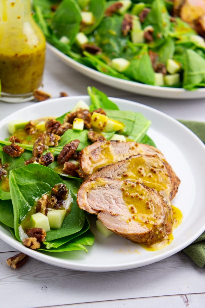 a small white plate with spinach, pecans, apples, and pork tenderloin on it drizzled with a yellow dressing in front of a carafe of the dressing and a large platter with the same salad.