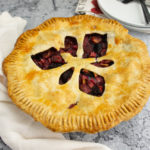 a rhubarb cherry pie on a grey stone table in front of a stack of plates