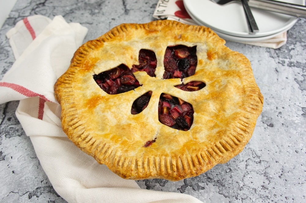 a rhubarb cherry pie on a grey stone table in front of a stack of plates
