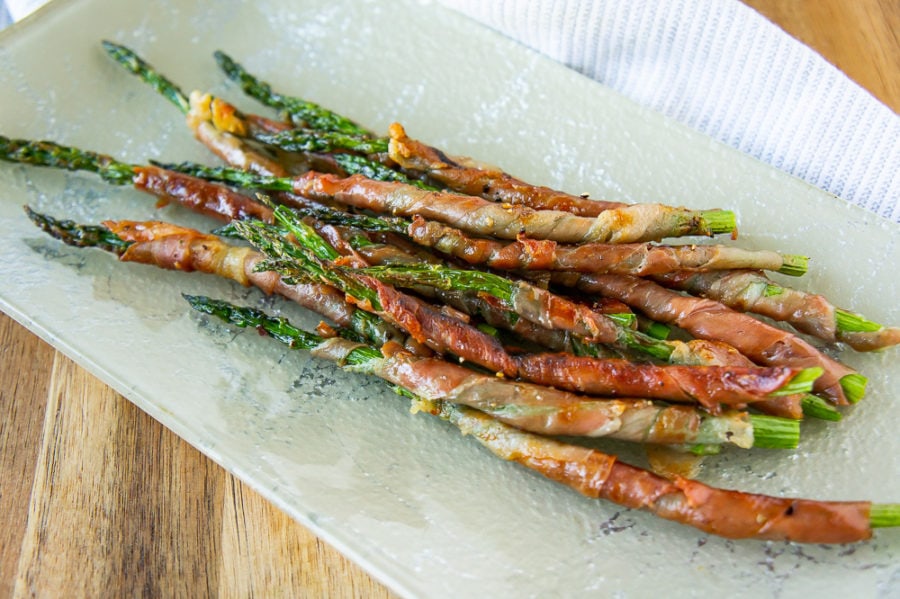 a platter of grilled prosciutto wrapped asparagus on a wooden cutting board