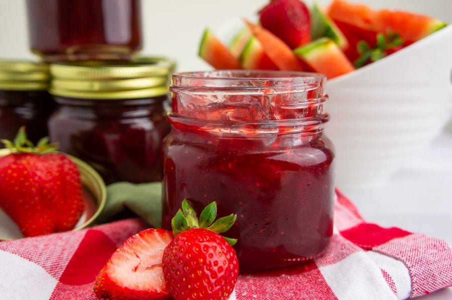 an open jar of strawberry watermelon jam on a red checked towel with sliced strawberries, a bowl of watermelon slices and extra jars of jam in the back. 