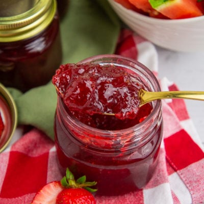 a jar of open strawberry watermelon jam with a gold spoon lifting jam out in front of other jars of jam and a bowl of watermelon on a white table