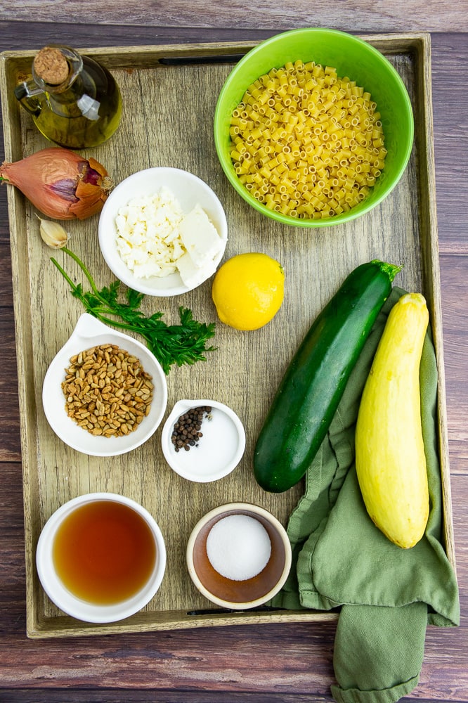 a serving tray with two summer squash, a bowl of uncooked pasta, a lemon, parsley, a shallot, olive oil, apple cider vinegar, and salt and pepper