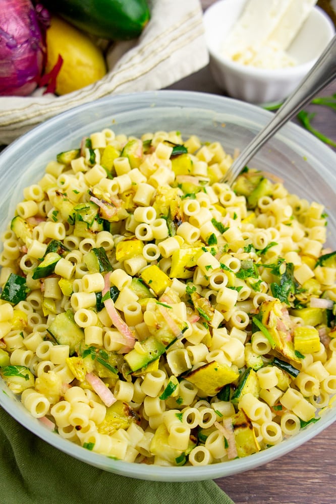 a large frosted bowl with summer squash pasta salad and a serving spoon on a brown wooden surface in front of a bowl of fresh vegetables.