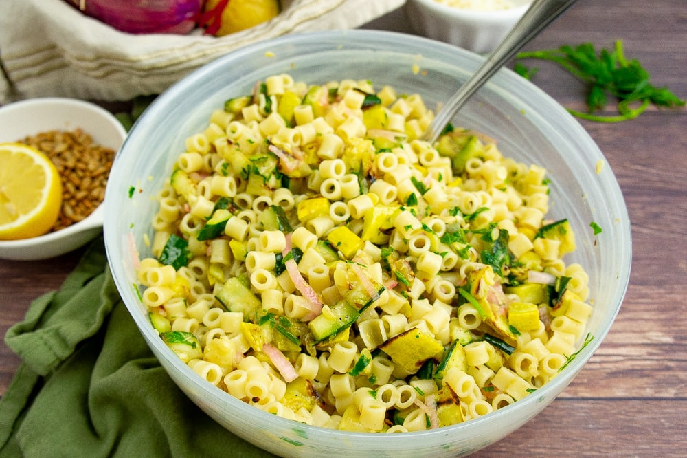 a frosted glass bowl with summer squash pasta salad on a wooden table beside a green linen towel