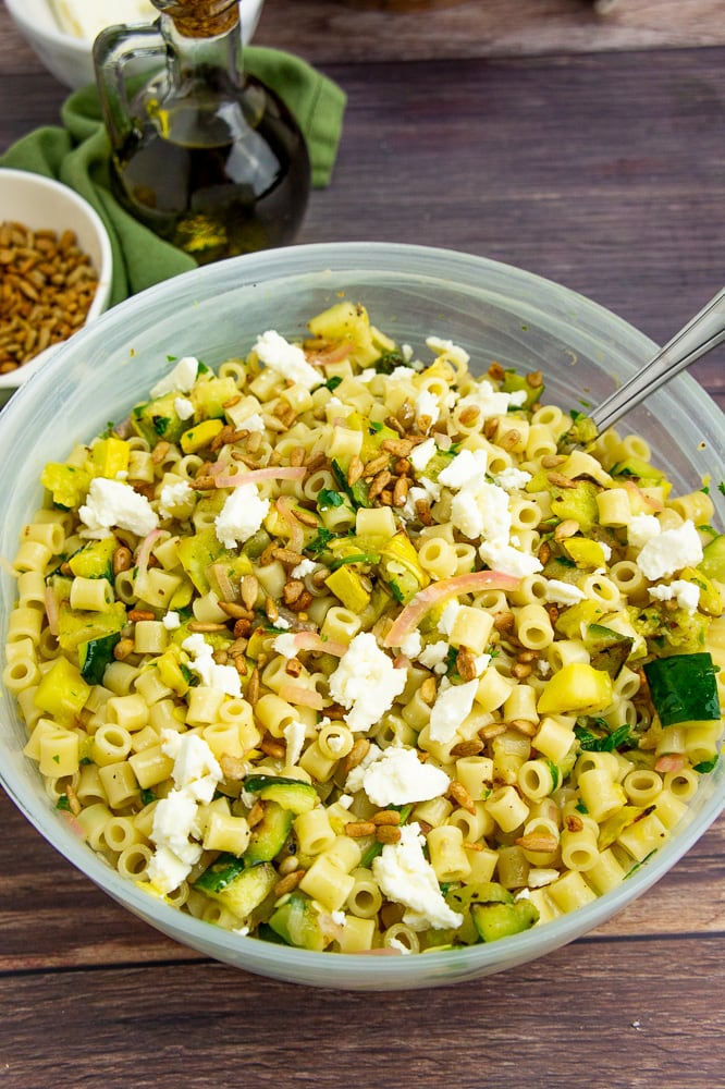 a bowl of pasta salad with a silver serving spoon topped with crumbled white cheese and sunflower seeds on a wooden table.