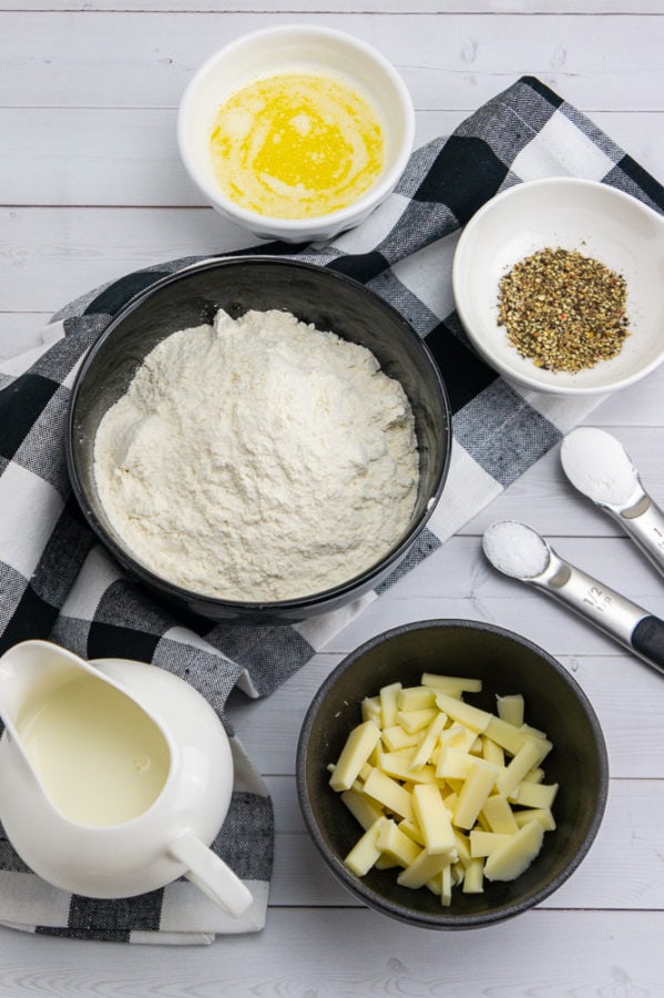 a bowl of flour with a bowl of asiago cheese with a carafe of milk, melted butter, and a bowl of black pepper on a black checked linen napkin