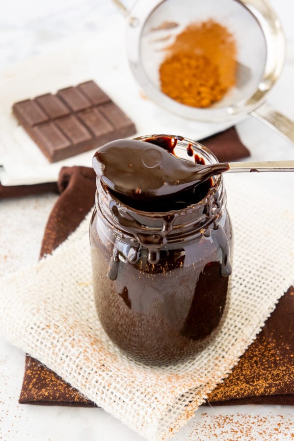 a jar of dark chocolate fudge sauce on a square of parchment paper with a sifter of cocoa powder