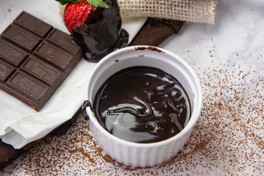 a small white ramekin of fudge sauce next to dark chocolate and strawberries