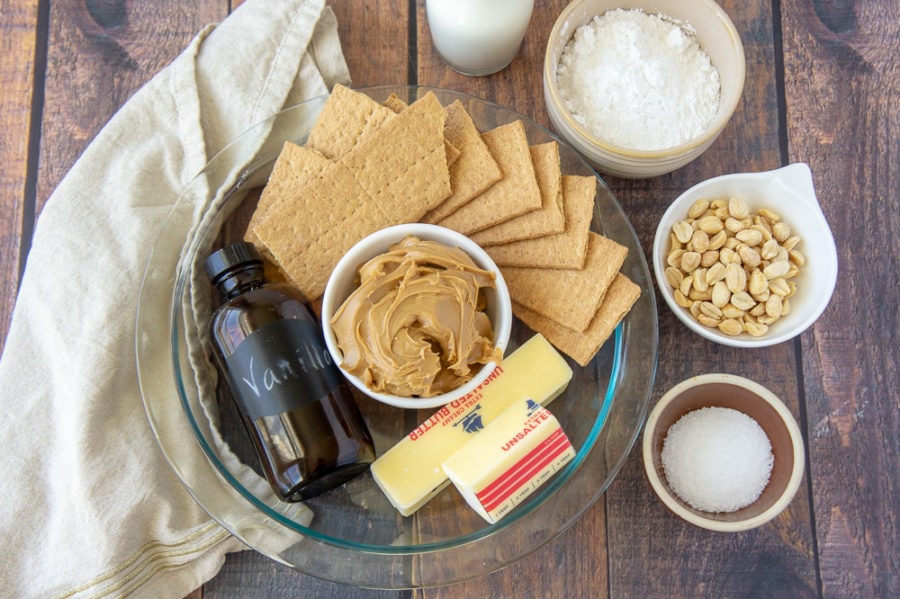 a glass pie plate with graham crackers, peanut butter, butter, and vanilla extract in there, with a bowl of peanuts, salt, and powdered sugar set beside it on a wooden table