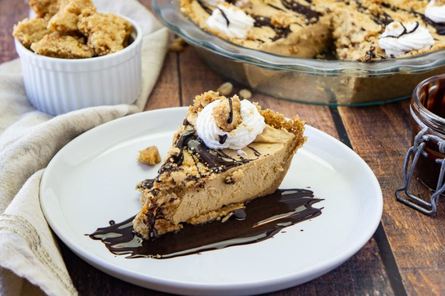 a slice of frozen peanut butter pie on a white plate with fudge sauce on a wooden table