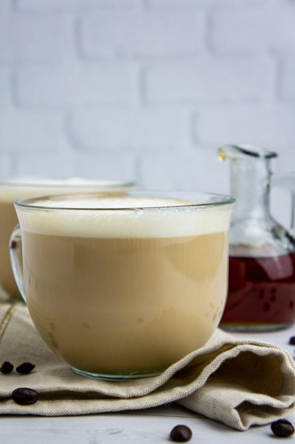 two cups of pale brown coffee drinks with a carafe of maple syrup on a white table