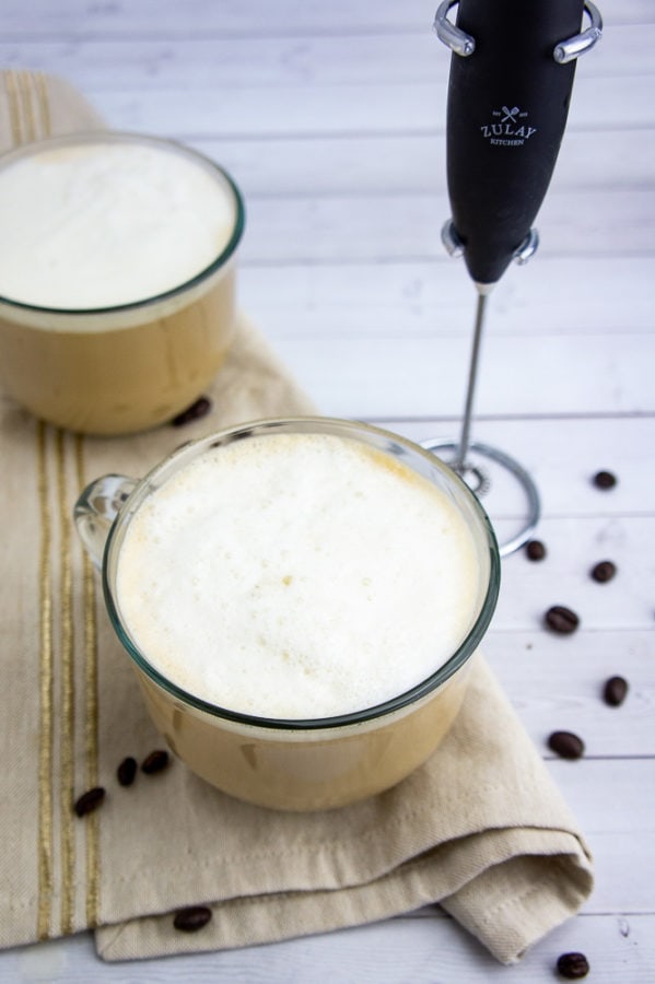 two lattes in glass cups on a brown napkin beside a milk frother