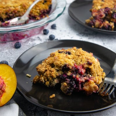 a black plate with plum blueberry cobbler and a fork on a stone surface in front of the full cobbler and a second plate