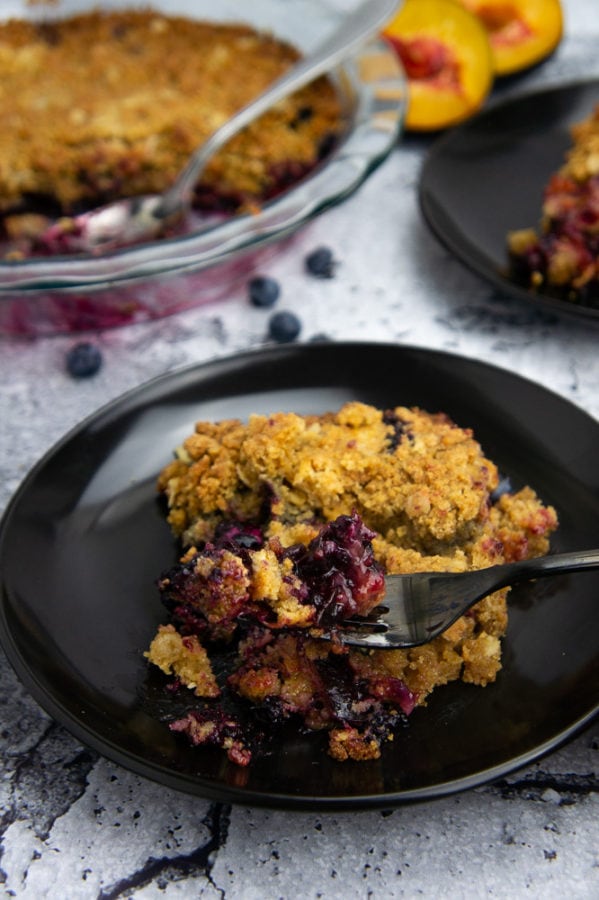 a black plate with plum and blueberry cobbler with a black fork on a stone surface.