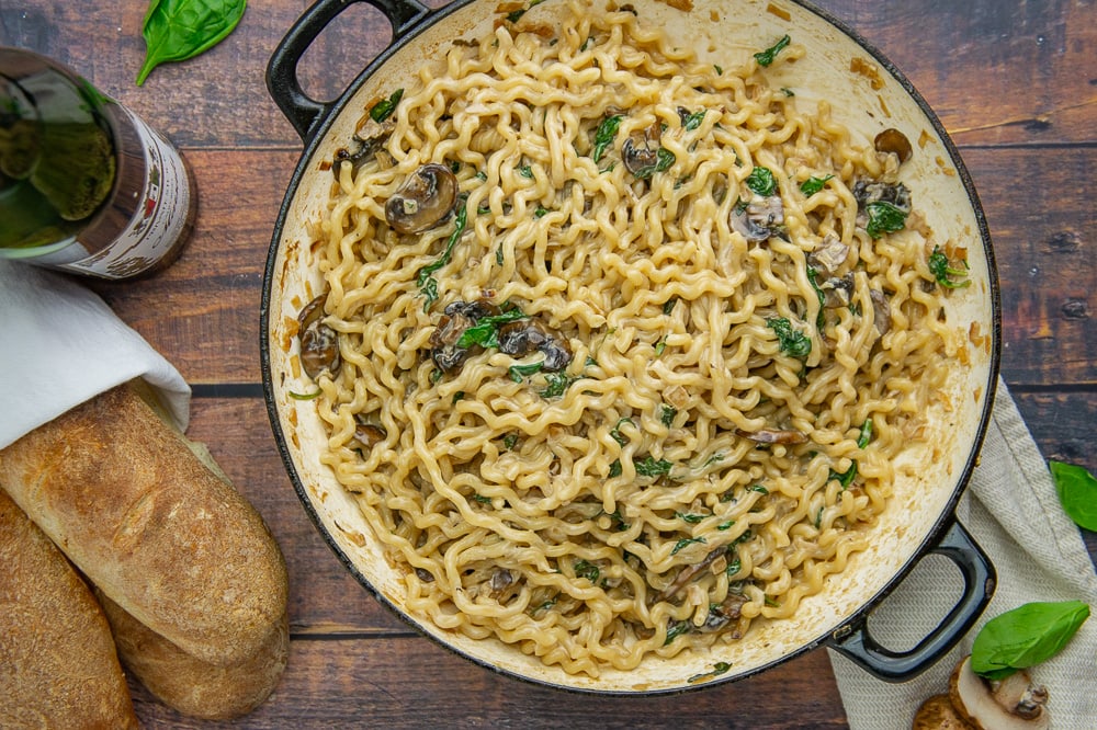 a large pot of wavy noddles with mushrooms and spinach in a creamy sauce beside some baguettes
