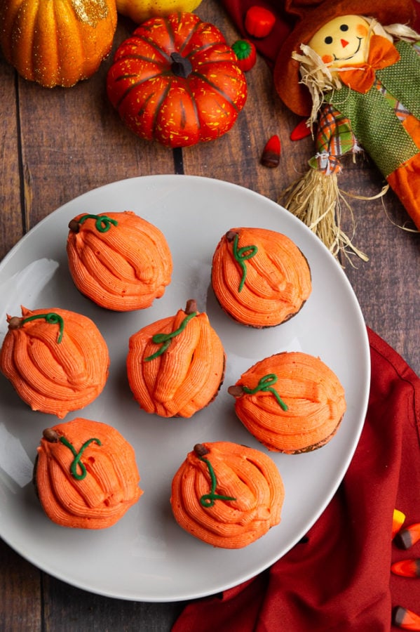 a white platter with pumpkin cupcakes frosted like orange pumpkins with a red napkin and a scarecrow on the table