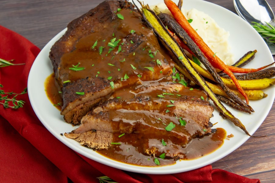 A white platter with sliced braised brisket in red wine sauce next to roasted carrots