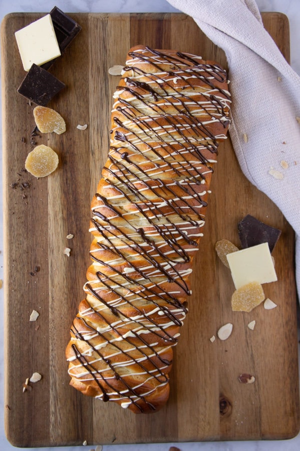 A whole chocolate drizzled braided chocolate loaf on a cutting board with bits of chocolate, almond, and ginger scattered around.