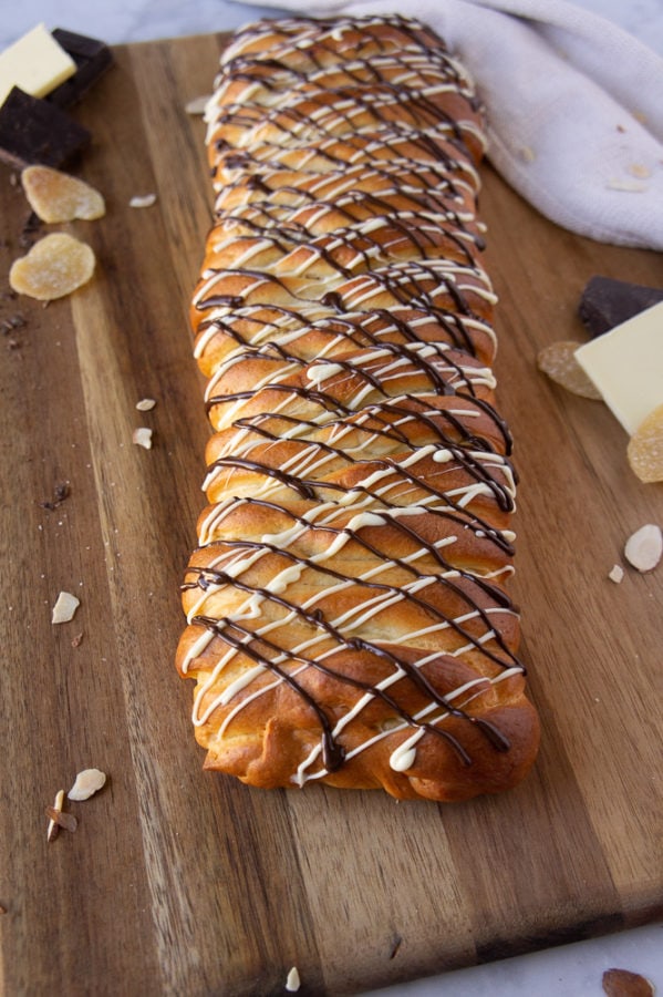 A braided loaf of bread drizzled with dark and white chocolate on a cutting board with bits of chocolate and crystallized ginger.