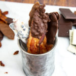 a metal cup with chocolate coated bacon standing in it on a white cutting board