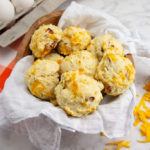 Ham and cheddar biscuits in a bowl beside cheddar cheese and eggs on a white table