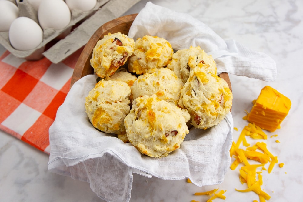 Ham and cheddar biscuits in a bowl beside cheddar cheese and eggs on a white table