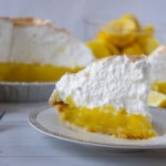 a slice of lemon meringue pie on a white plate with lemons and more pie behind it