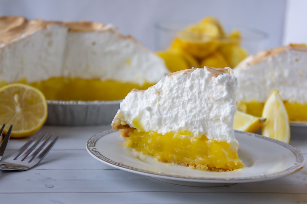 a slice of lemon meringue pie on a white plate with lemons and more pie behind it