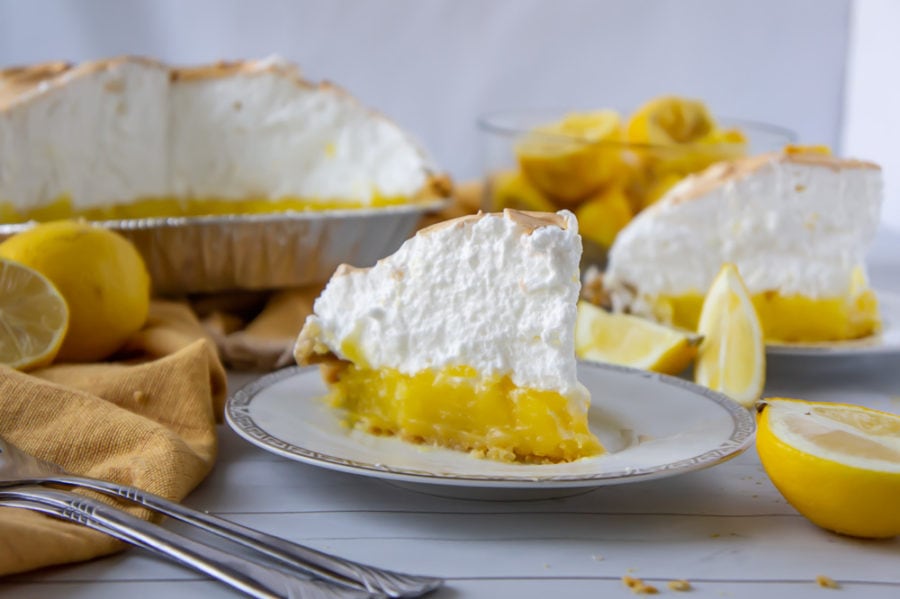 a slice of lemon meringue pie on a white plate with lemons and more pie behind it