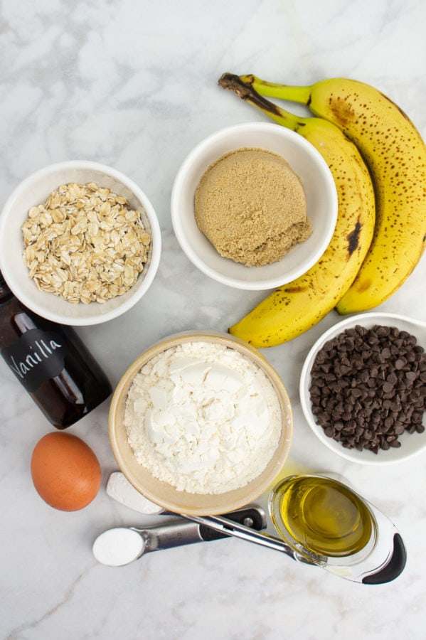 a bowl of white wheat flour beside bowls of oats, sugar, chocolate chips, and two whole bananas