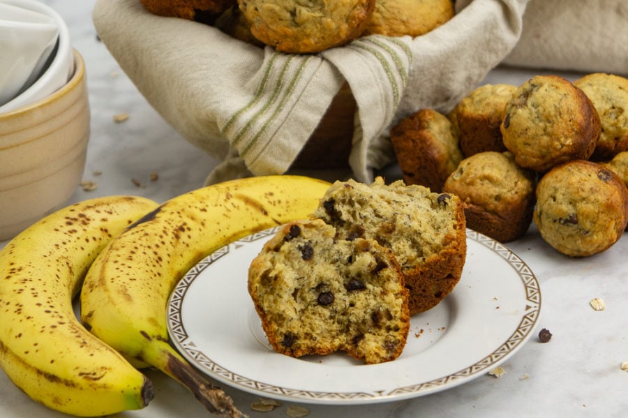 a banana oat muffin open on a white plate beside bananas