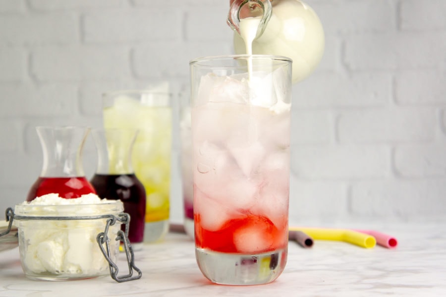 cream being poured into a tall glass with red syrup, ice and club soda