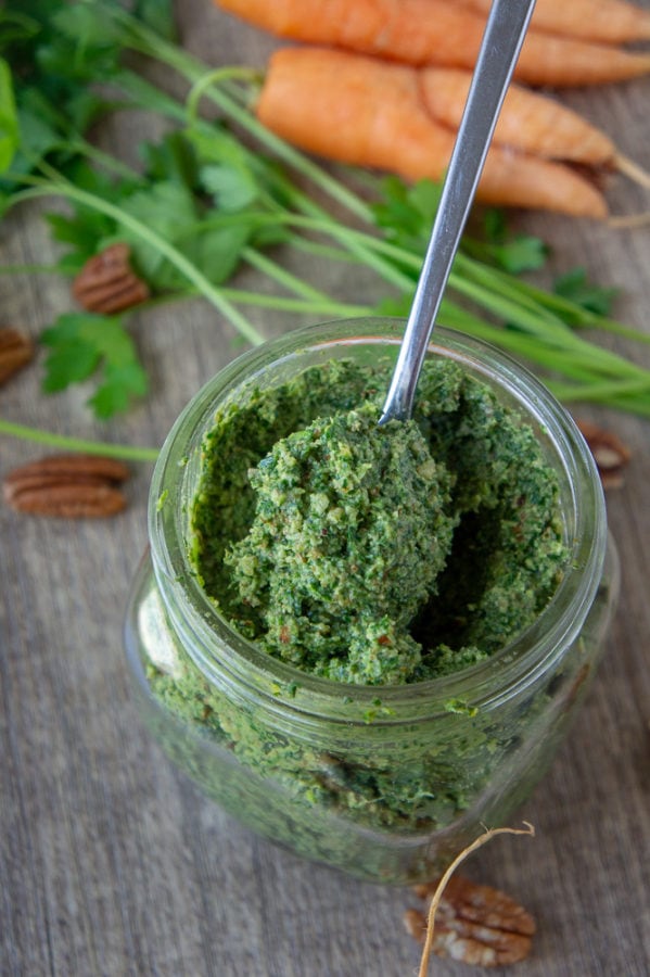 a jar of carrot top pesto with a spoon in it in front of some carrots and parsley