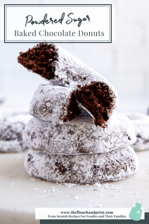 a stack of chocolate baked donuts coated in powdered sugar with one cut open on top